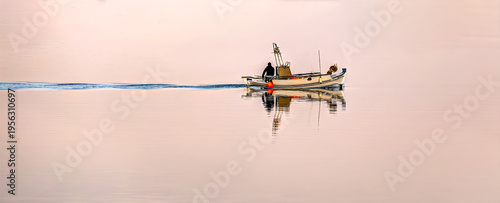 Foto Lemnos, Fisher on a small boat creating a ripple wake on the mirror-like water, reflecting the soft sunrise colors