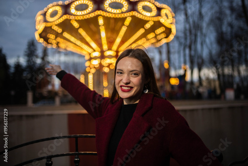 A woman in an amusement park with a carousel in the background is happy and smiling in the evening