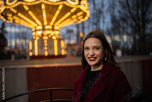 A woman in an amusement park with a carousel in the background is happy and smiling in the evening
