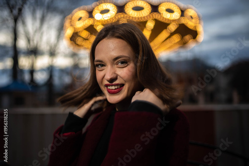 A woman in an amusement park with a carousel in the background is happy and smiling in the evening