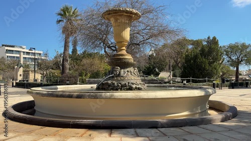 Ancient stone fountain with cherub sculptures and large decorative urn in a public park on Malta. Elegant architectural monument surrounded by palms and greenery under a clear blue sky.