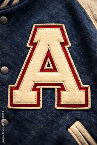 Close-up of a cream-colored felt letter A with red trim, stitched onto a navy blue varsity jacket.