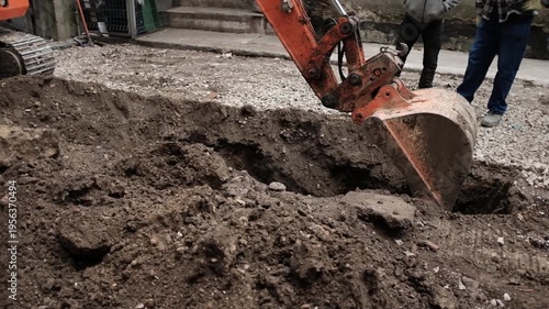 Close-up video of a heavy-duty orange hydraulic excavator bucket digging a deep narrow trench in dark, moist soil at an urban construction site. The video shows the precise mechanical movement.