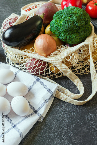 Nutrient rich fresh vegetables in a reusable string bag and white eggs resting on a plaid towel on a kitchen surface
