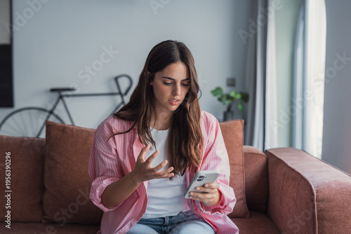 Obraz na plátně Young woman on a couch reacting with frustration and confusion while looking at her mobile phone in a living room, conveying stress, annoyance, and digital communication problems