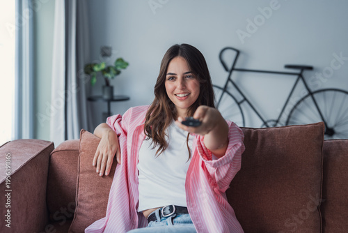 Smiling young woman lounges on a sofa in a bright living room, pointing a TV remote toward the screen and enjoying a relaxed moment at home that conveys comfort, leisure and modern lifestyle.