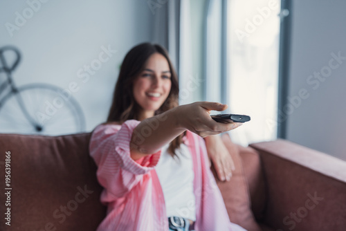 Young woman lounges on a couch in a bright living room, smiling and holding a TV remote as she relaxes and watches television, enjoying leisure time at home.