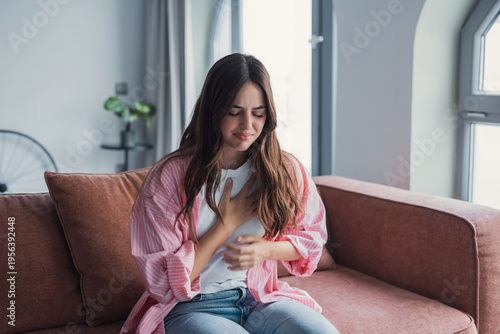 Young woman sits on a living room couch clutching her chest in visible discomfort, suggesting heartburn, indigestion or anxiety-related chest pain in a domestic setting.