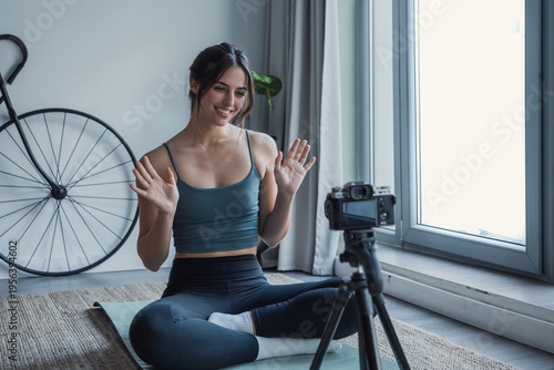 Photos A smiling young woman records a home yoga session for a fitness vlog, seated on a mat and engaging viewers with a friendly wave and professional camera setup by the window