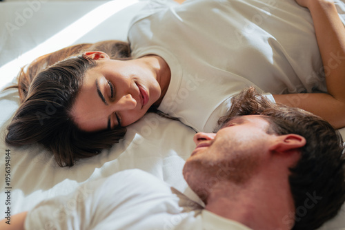 A young couple lies facing each other on a white bed, exchanging warm smiles and gentle eye contact, conveying intimacy, comfort and peaceful morning togetherness in a bright bedroom.