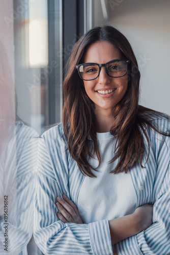 Free Download Smiling young woman wearing glasses and a striped shirt standing by a window, casual professional portrait conveying confidence and approachability - GFXLoad