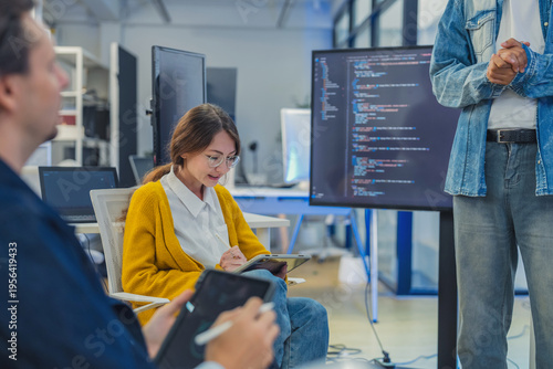 collaborative female AI developer in yellow sweater and glasses discusses code and neural network diagrams on large monitor while taking notes on tablet in modern tech office, with team members listen