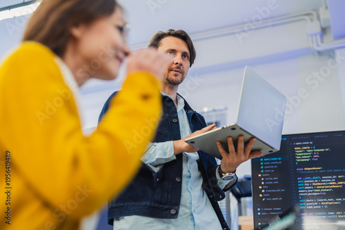 collaborative female AI developer in yellow sweater and glasses discusses code and neural network diagrams on large monitor while taking notes on tablet in modern tech office, with team members listen