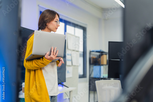 confident female AI team leader in yellow cardigan and glasses presents neural network architecture and code on large monitor while explaining to seated developers with laptops and tablets in office