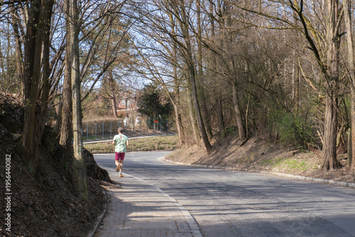 Man running on a paved path beside a road through a wooded area in early spring