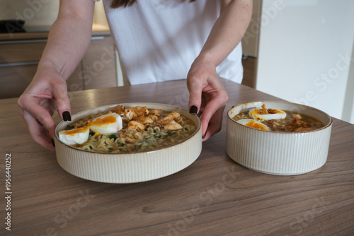 Hands serving delicious ramen noodles with boiled eggs and chicken on a wooden table