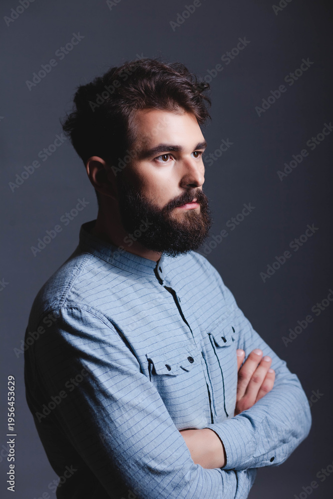 Obraz premium Studio portrait of a serious, confident young bearded man standing with arms crossed, looking in profile to the right against a solid dark grey background with copy space.
