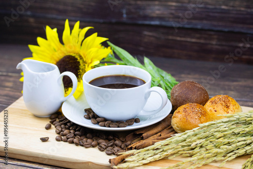 Still life coffee scene with cup of black coffee, bread, coffee beans, sunflower and cinnamon on wooden background, warm cozy cafe