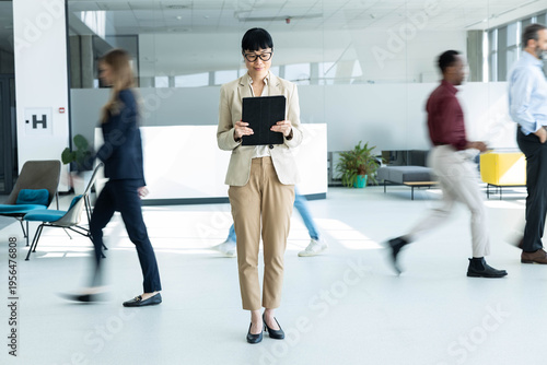 Canvas Print Businesswoman standing calmly, looking at tablet while blurred colleagues walk q