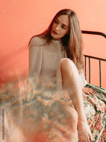 Stylish caucasian woman in boudoir dress sitting on bed with warm background and artistic light effects.