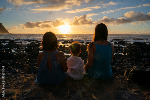 Silhouettes of two adults and a child on rocky shore at sunset. Golden sun dips behind distant hills, casting warm glow. Calm ocean reflects light, blending with sky's soft hues