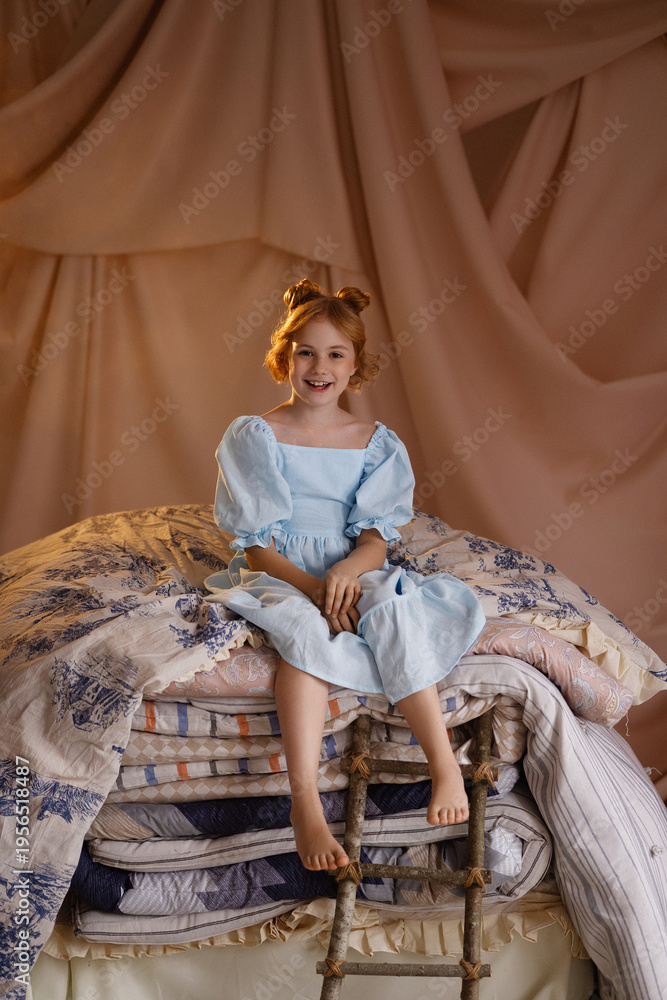 Obraz premium Smiling Caucasian girl in blue dress sitting on stacked mattresses like princess and the pea, front view, look at camera, studio with beige drapery. Concept of fairy tale, childhood imagination.