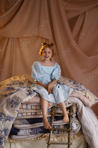 Smiling Caucasian girl in blue dress sitting on stacked mattresses like princess and the pea, front view, look at camera, studio with beige drapery. Concept of fairy tale, childhood imagination.