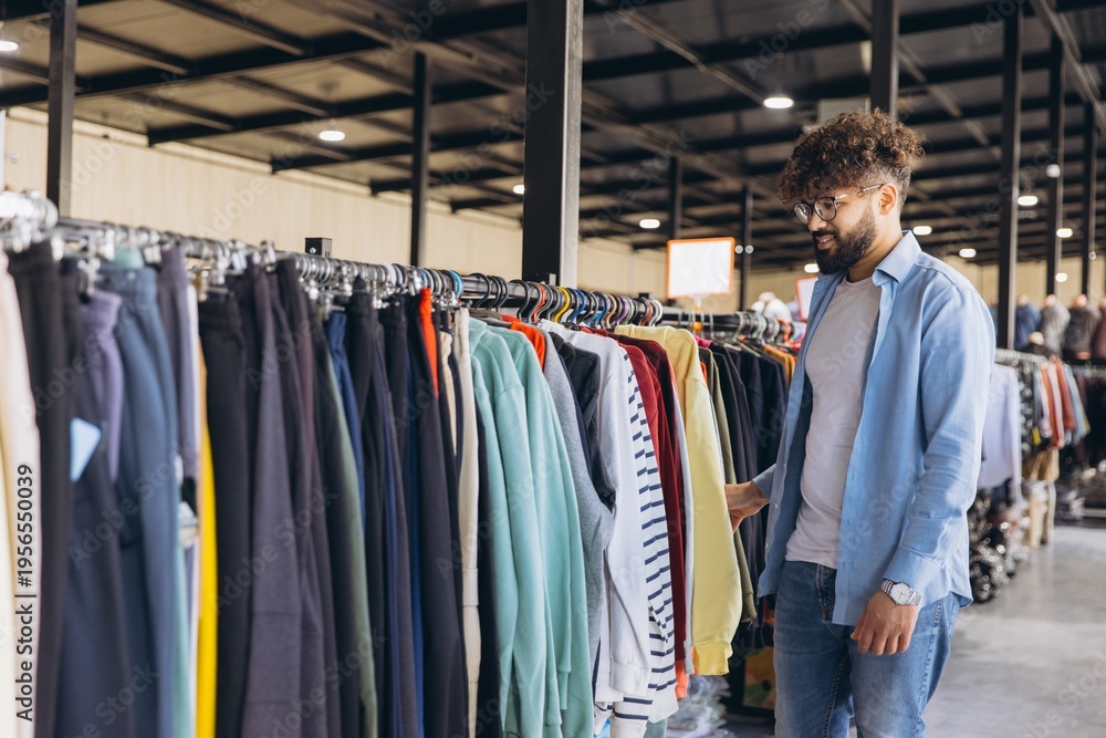 Obraz premium Arab man browsing clothing rail in a store, choosing new apparel, enjoying a shopping experience