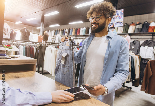 Fotografi Smiling Arab man purchasing jeans using contactless payment with a smartphone in