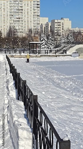 An adult in a yellow jacket is cross-country skiing across a frozen snow-covered pond in a city park on a sunny winter day.