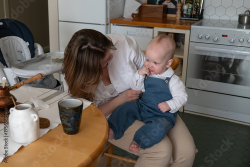 Mother sits with baby near table in kitchen interior with natural light and home setting, everyday interaction and care moment in domestic environment. Family bonding, motherhood, parenting