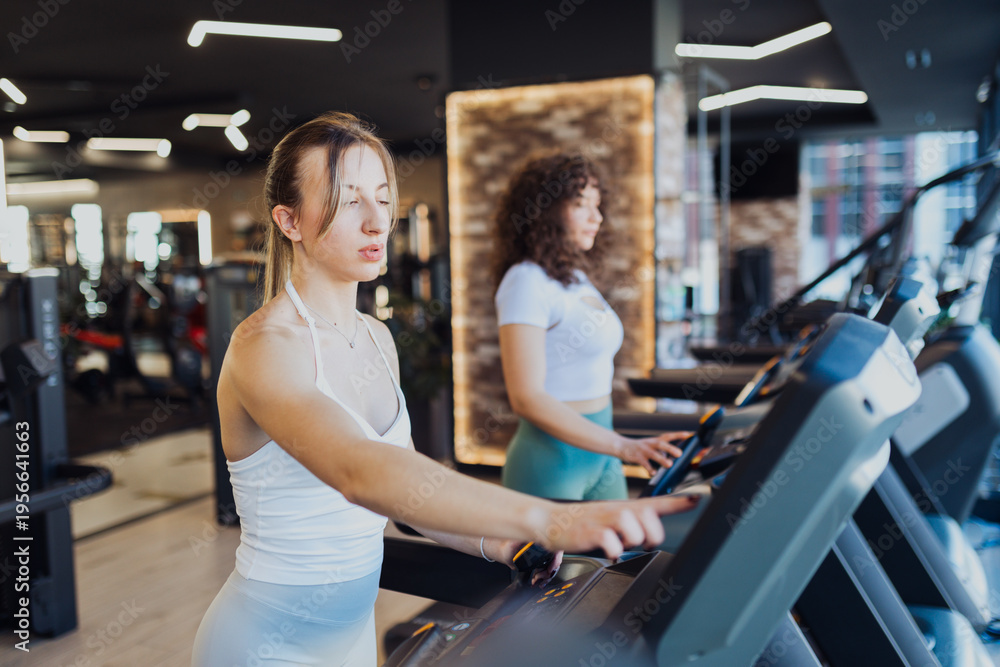 Fototapeta premium Two young women preparing to run on treadmills in a modern gym. 