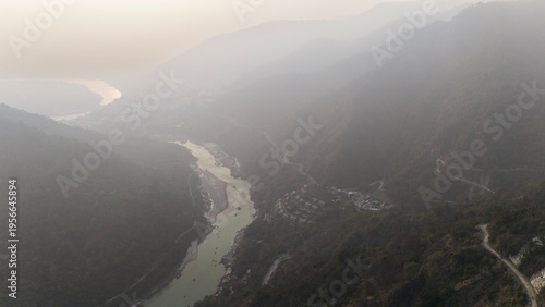Fotografi Aerial landscape view from Kuee village in Tehri Garhwal, Uttarakhand, showing misty mountains and forested hills under clouds, with the Ganga River flowing below on a winter evening