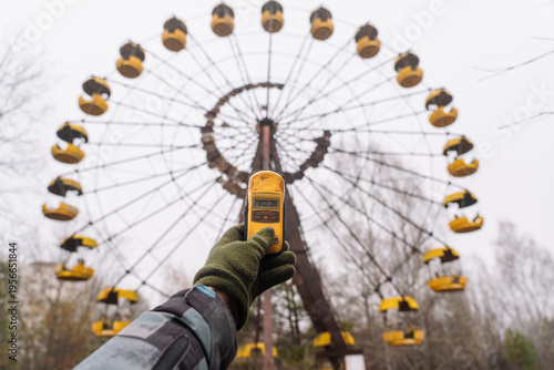 A man with a dosimeter in hand near ferris wheel. Abandoned amusement park in Pripyat city. Chernobyl Exclusion Zone.