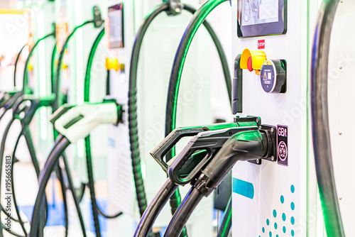 Row of white electric vehicle chargers with black cords, digital displays green and blue, even lighting casting soft shadows to the right, on a textured white wall Modern, functional style No hum