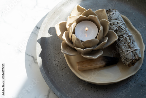 Lotus candle holder with burning tea light, white sage smudge stick and palo santo on concrete plate over white marble. Zen spiritual cleansing concept. 
