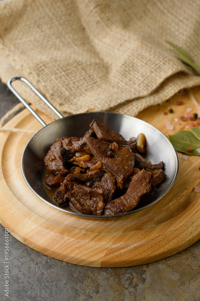 Fototapeta premium Braised Beef Strips with Onion in Metal Pan on Wooden Board with Bay Leaves, Pink Salt and Burlap on Stone Background