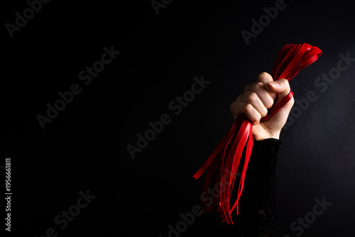 Human hand holding red flogger whip against dark background, studio lighting highlighting leather straps, texture, and grip.