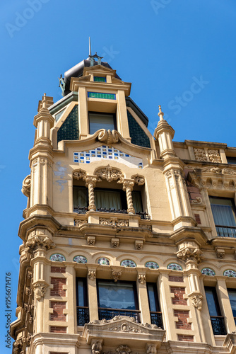 Detailed view of a historic building in Gijon, Asturias, featuring intricate stone carvings, colorful mosaic tiles, and a traditional spire against a clear blue sky.