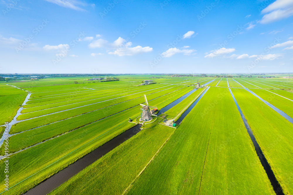 Fototapeta premium Aerial view of traditional Dutch windmills by water canals in a green field. Historic heritage site in Netherlands under a blue sky. Rural landscape with classic architecture and agriculture.