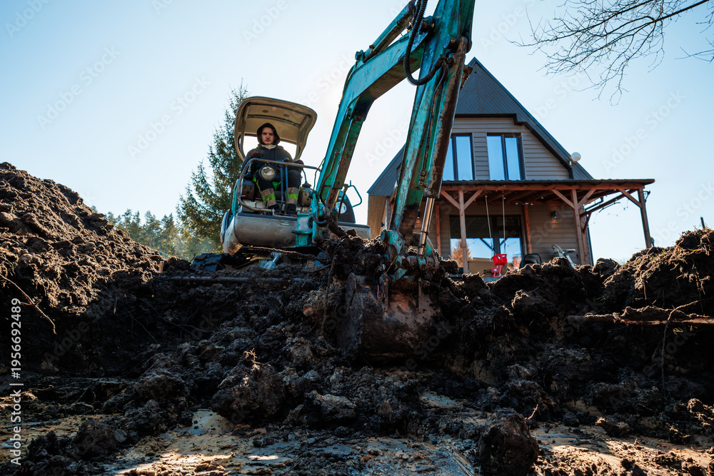 Fototapeta premium A person operates a teal mini excavator, digging wet soil before an A frame cabin with large vertical windows and a timber porch, conifers border the rural site.
