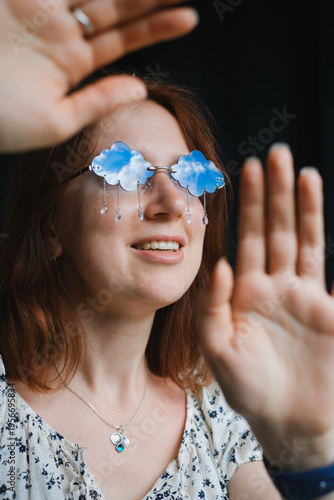 Smiling redhead woman wears creative cloud-shaped sunglasses reflecting a blue sky. A dreamy, optimistic concept for creativity, imagination, and positive thinking.