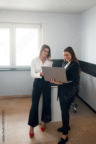 Two professional women standing with laptop and discussing company performance in modern office.