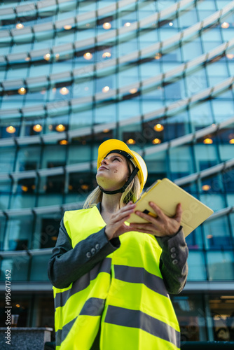 Photography Woman architect wearing hardhat and safety vest holding tablet, looking up at bu