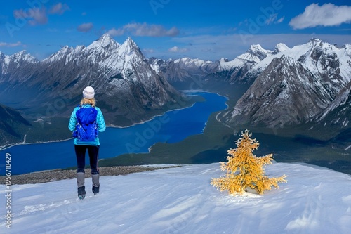 A lone hiker stands on a snowy mountain summit overlooking a dramatic alpine landscape with jagged peaks and a deep blue glacial lake below with golden larch in snow 