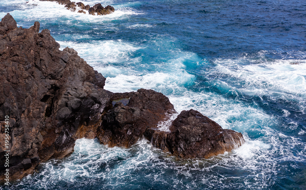 Naklejka premium Basalt cliff shoreline. Surf crashes against jagged volcanic rocks at Porto Moniz, Madeira, Portugal. White sea foam and wave spray contrast with deep blue Atlantic water.