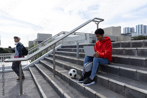 Diverse teenage males sitting and walking on steps with laptop phone and soccer ball, copy space