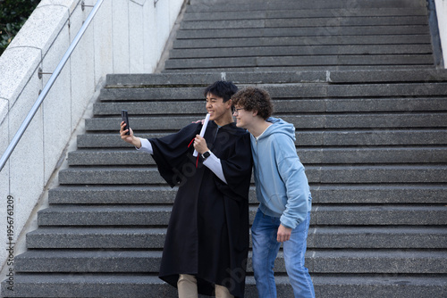 Two males in late teens taking selfie, graduate wearing black gown holding diploma on steps
