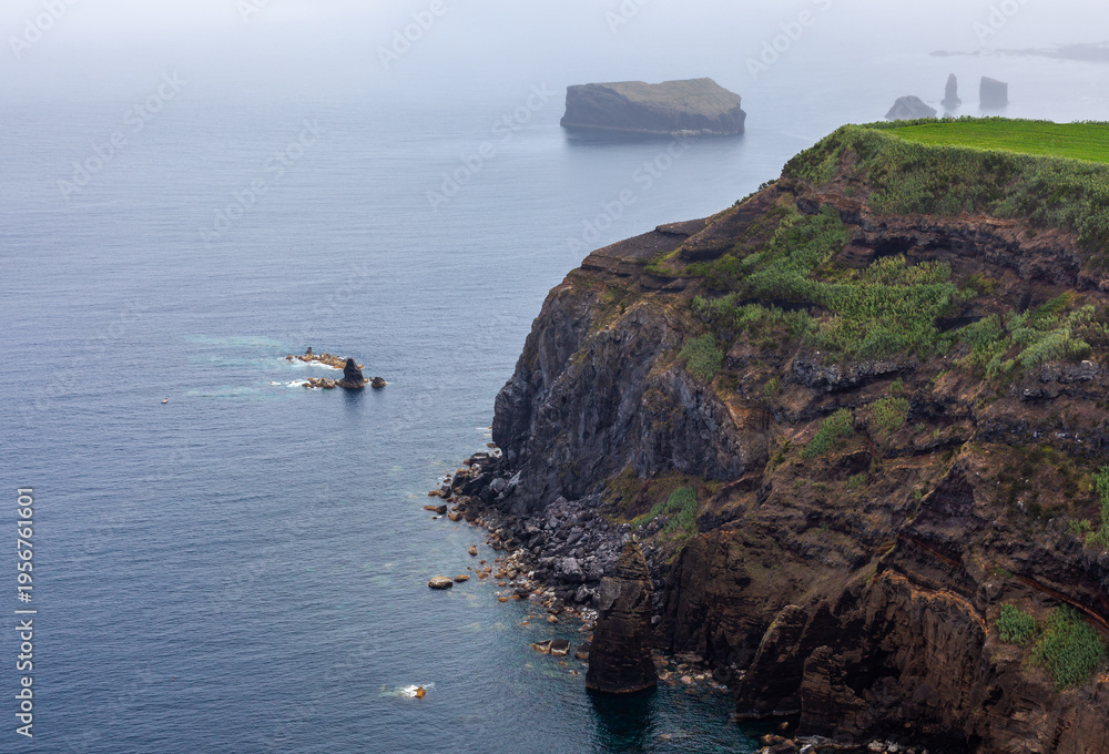 Obraz premium Rugged volcanic cliffs along the Atlantic coast of San Miguel Island, Azores, Portugal. Rocky shoreline with green vegetation. Small islands visible in the distance.