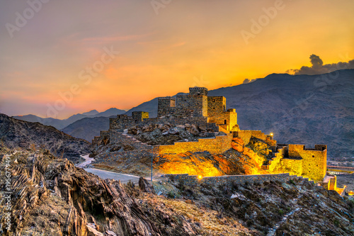 A scenic view of the ancient stone structures of Thee Ain Heritage Village illuminated at sunset against the mountains in Al Bahah, Saudi Arabia.